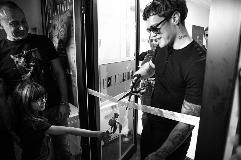 Foto di inaugurazione di una pasticceria con ragazzo con occhiali da sole, tatuaggi e maglietta nera taglia un nastro davanti a una bambina e un signore